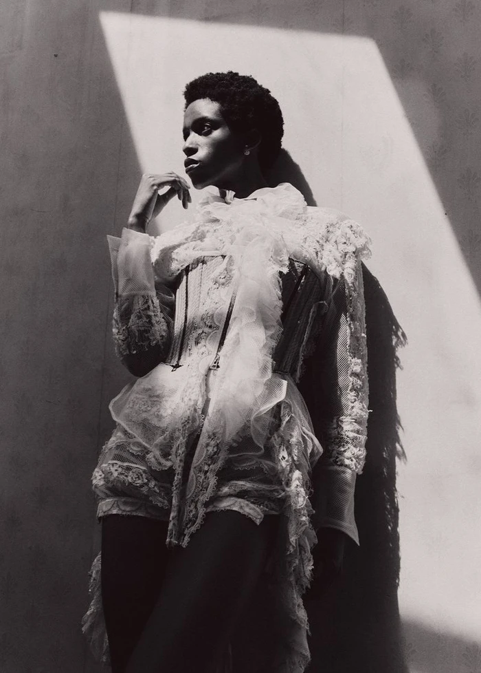 Black female model in an intricate white ruffled lace corset ensemble posing against a wall in dramatic diagonal sunlight.