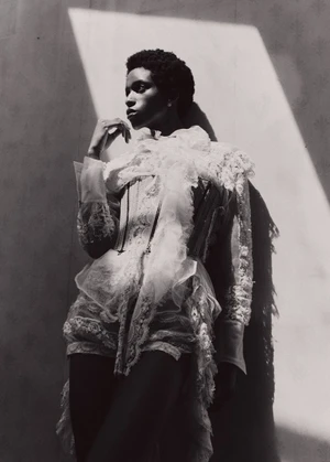 Black female model in an intricate white ruffled lace corset ensemble posing against a wall in dramatic diagonal sunlight.