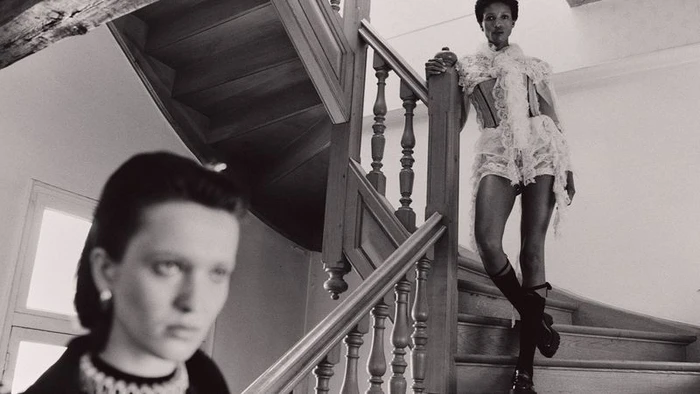 Black and white fashion shot of a model in a lace corset outfit on a wooden staircase with another model blurred in the foreground.