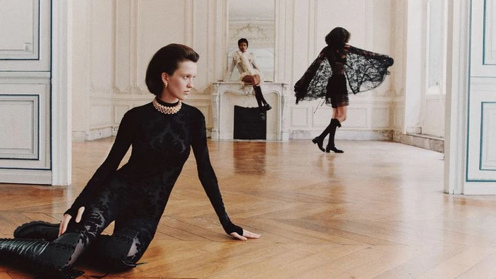 Model in a black patterned catsuit reclining on a wooden parquet floor alongside two models in a spacious room.
