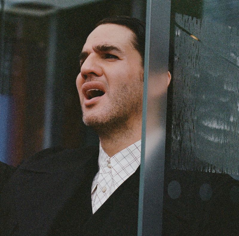 Close-up of adult male with open mouth, wearing a black sweater and white shirt, positioned behind a glass structure indoors.