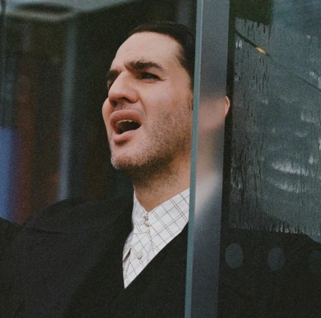 Close-up of adult male with open mouth, wearing a black sweater and white shirt, positioned behind a glass structure indoors.