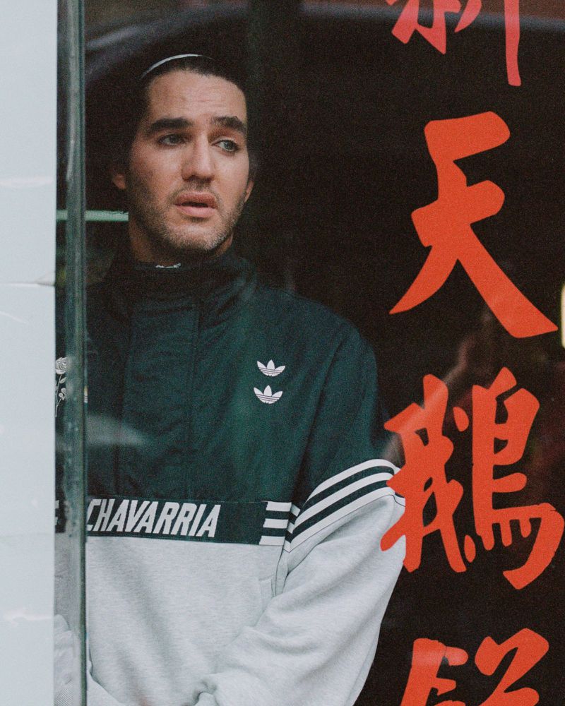 Portrait of a young adult male wearing a green and white jacket, gazing off-camera with a Chinese text poster in the background.