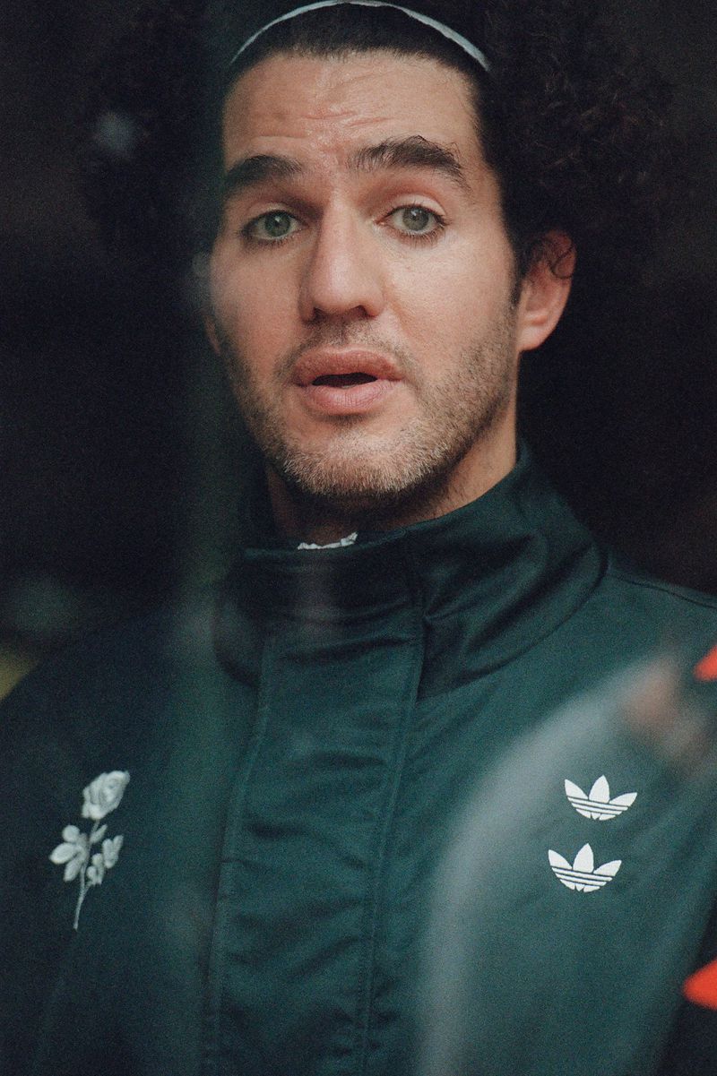 Close-up portrait of an adult male with curly hair wearing a white headband and a green Adidas jacket, looking slightly off-camera.