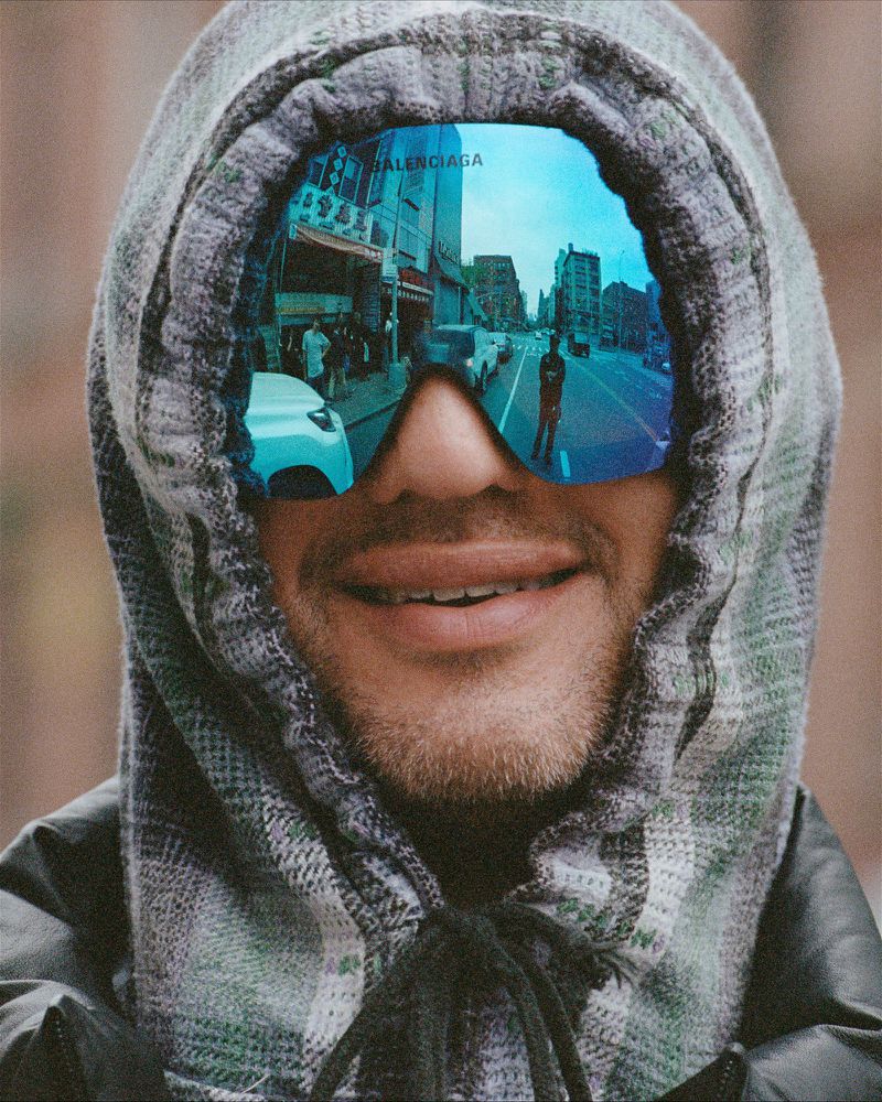 Close-up of a smiling man in a hooded plaid jacket, sunglasses reflecting a city street with cars and pedestrians.