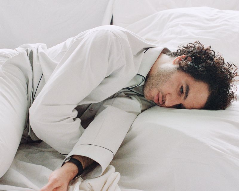 Man with curly hair rests on a white bed, wearing grey pajamas with black trim. Indoor scene with natural lighting, close-up portrait.
