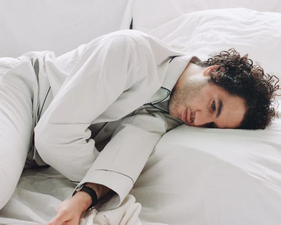 Man with curly hair rests on a white bed, wearing grey pajamas with black trim. Indoor scene with natural lighting, close-up portrait.