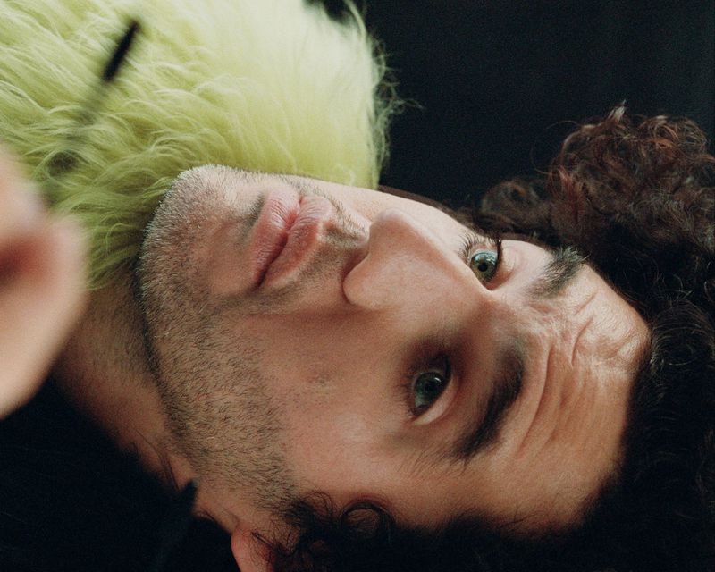 Portrait of an adult man with dark hair and beard laying down wearing a yellow fur-like collar, looking directly at the viewer.