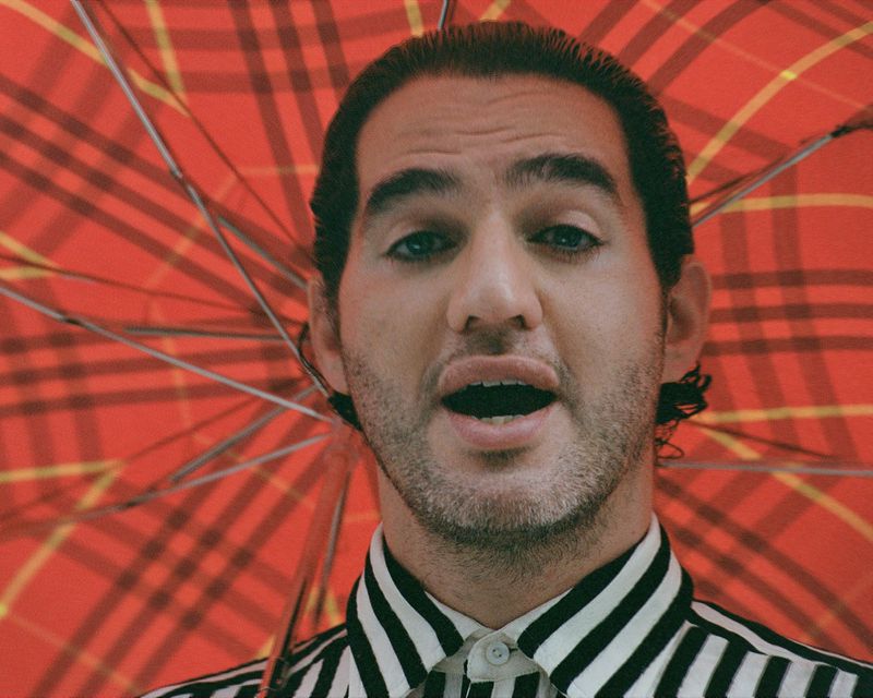 Close-up portrait of a man with dark hair and a striped shirt, standing under a red plaid umbrella, looking directly at the camera.