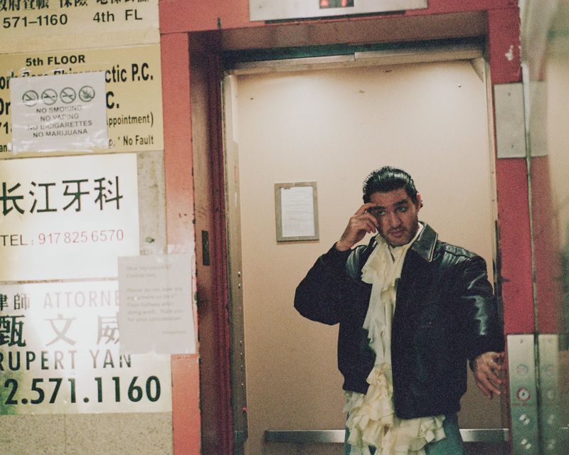 A man stands in an elevator with posters on the wall, wearing a black jacket and jeans with a ruffled white shirt.
