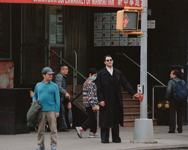 Man in black coat presses crosswalk button on city street with other pedestrians and a 'do not walk' sign in Manhattan.
