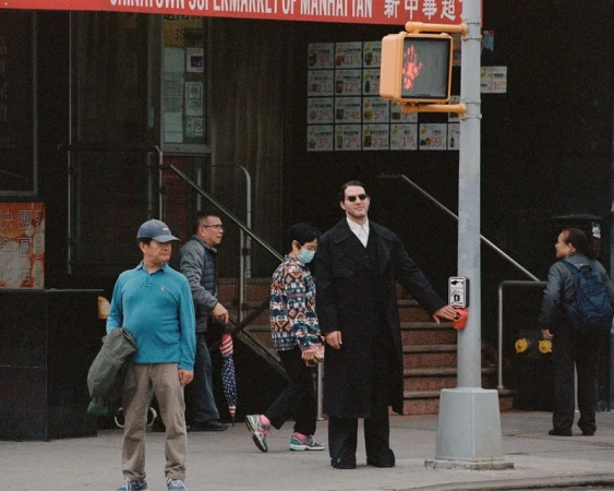 Man in black coat presses crosswalk button on city street with other pedestrians and a 'do not walk' sign in Manhattan.