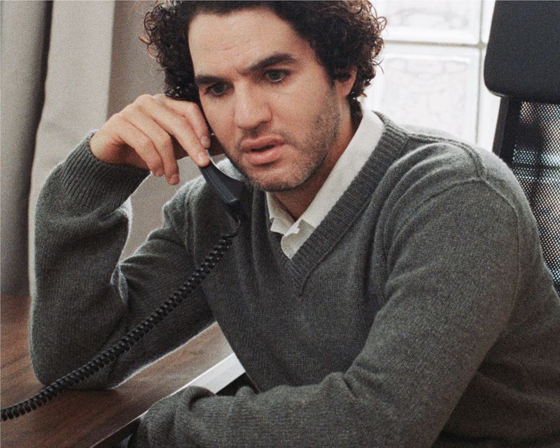 Man in grey sweater on phone at desk. Indoor scene shows adult male with curly hair talking on a landline telephone in natural light.