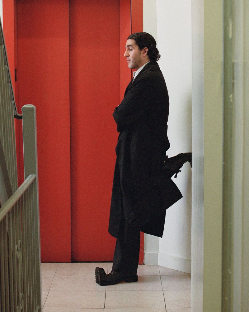Man in a black overcoat poses in an indoor hallway, standing against a wall with a red door and stair railing visible. 