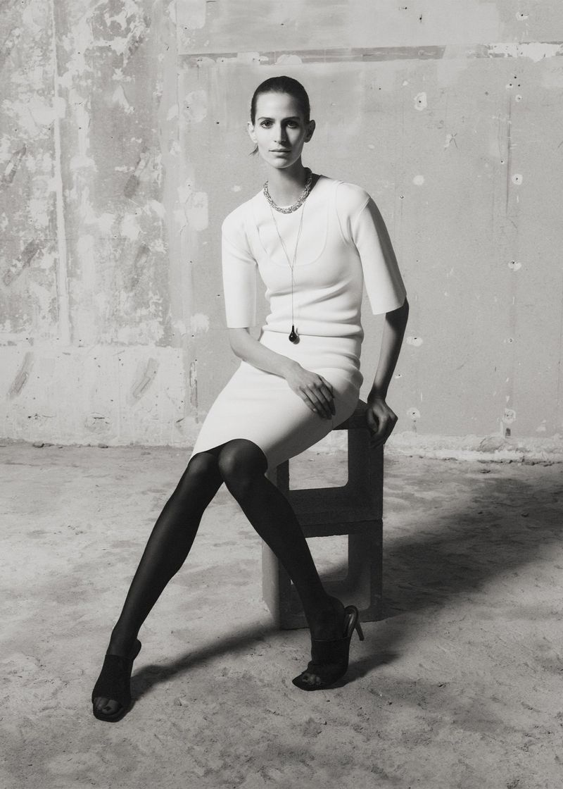 A monochrome fashion shot of a slender adult woman sitting on a block stool, wearing a dress and tights with heels, against a textured wall.