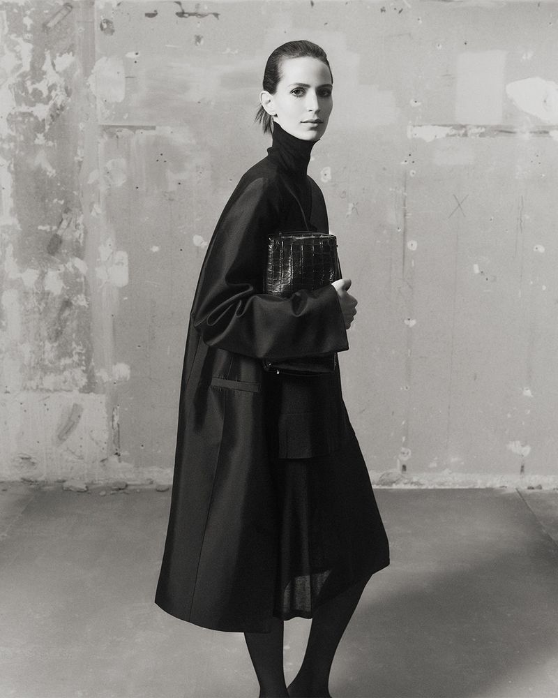 Jeanne Cadieu holding a dark structured handbag wearing a black coat against a weathered wall for Totême