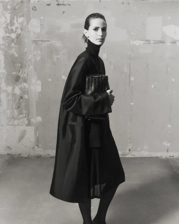 Jeanne Cadieu holding a dark structured handbag wearing a black coat against a weathered wall for Totême
