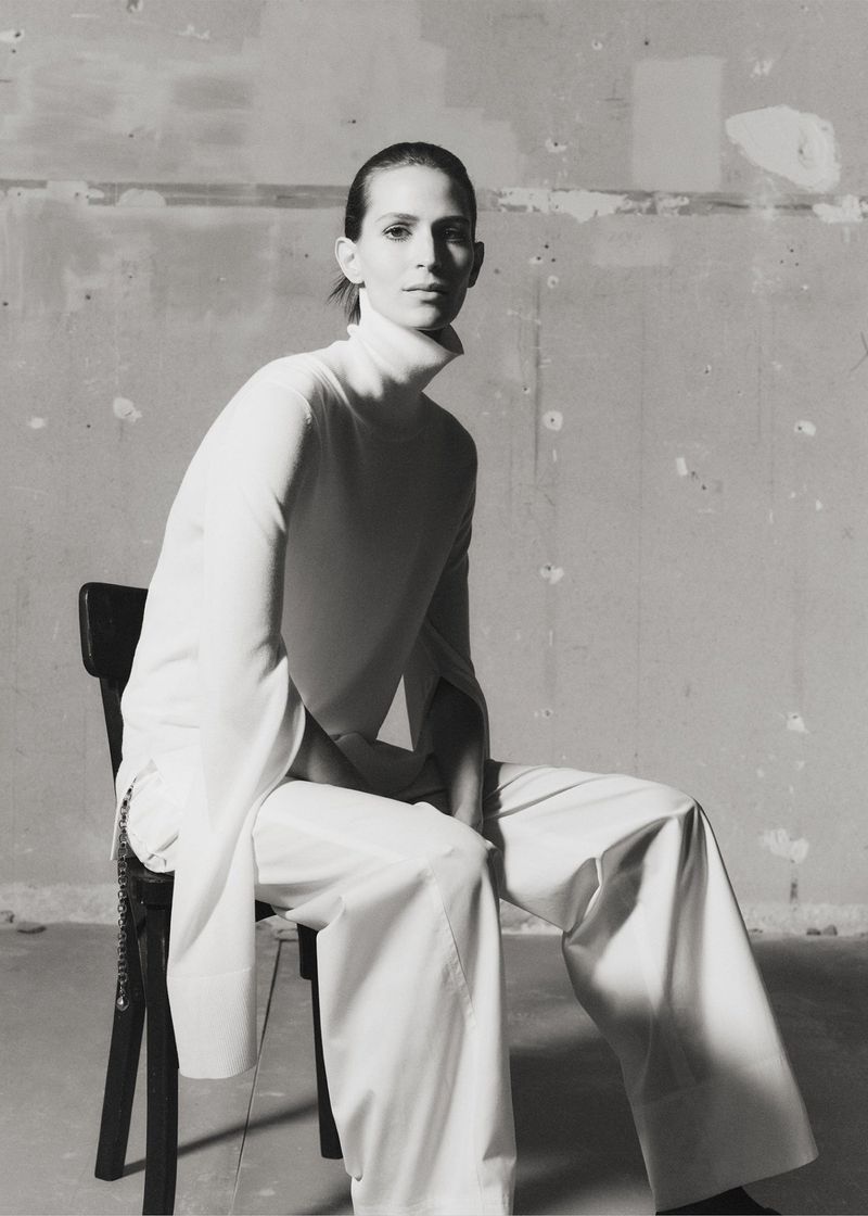 Black and white portrait of a slender, fair-skinned adult female sitting on a black chair in a minimalist studio wearing wide-leg white pants.
