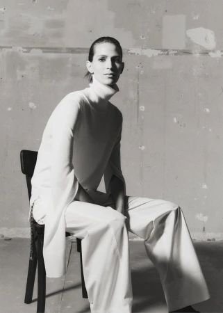 Black and white portrait of a slender, fair-skinned adult female sitting on a black chair in a minimalist studio wearing wide-leg white pants.