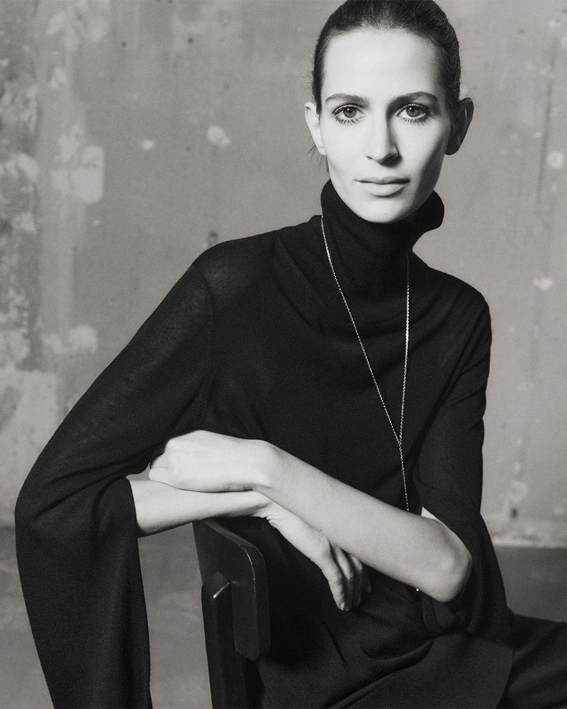 Elegant portrait of a seated adult woman with dark hair wearing a black turtleneck, captured in a monochrome, minimalist setting.