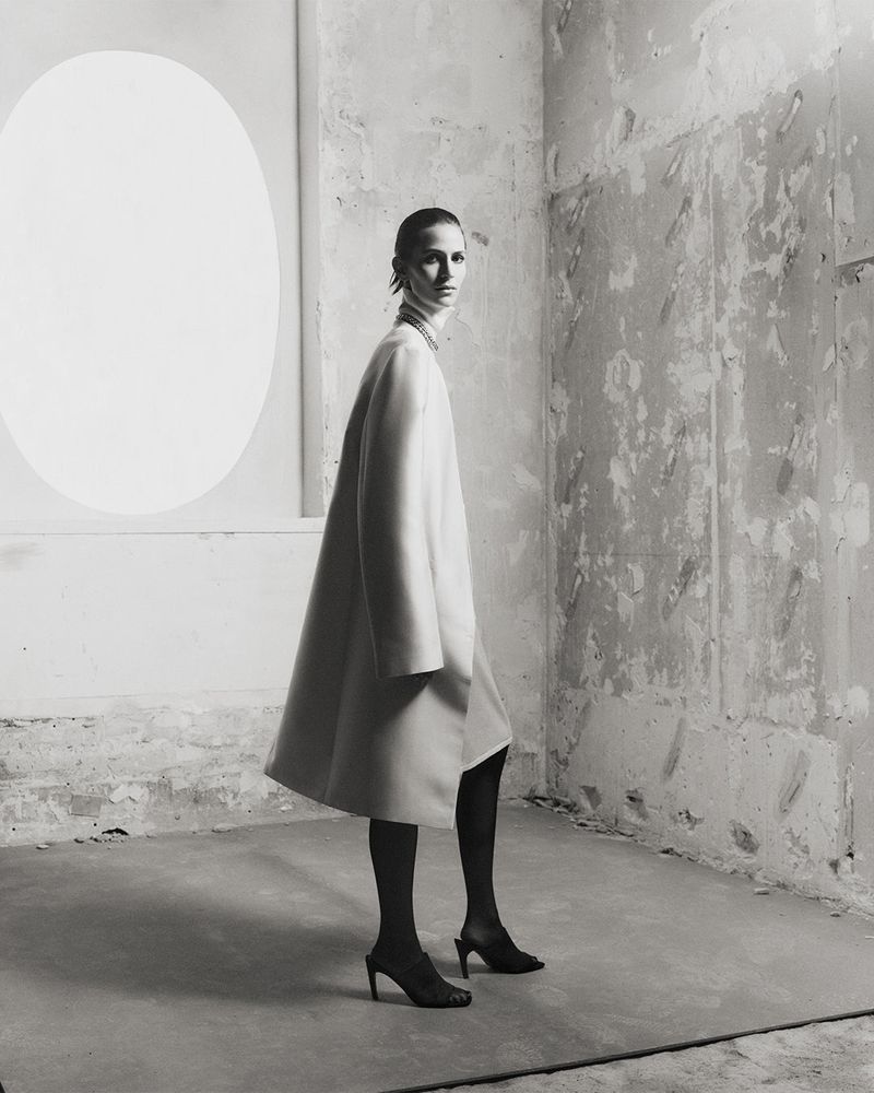 A woman in a long cream coat and black tights poses in a sparsely decorated room with a distressed wall and a white oval shape.