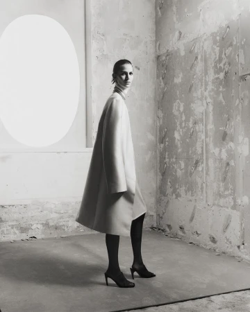 A woman in a long cream coat and black tights poses in a sparsely decorated room with a distressed wall and a white oval shape.