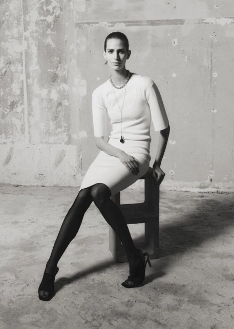 Jeanne Cadieu seated on a stool wearing a white knit dress and black hights against a distressed concrete wall