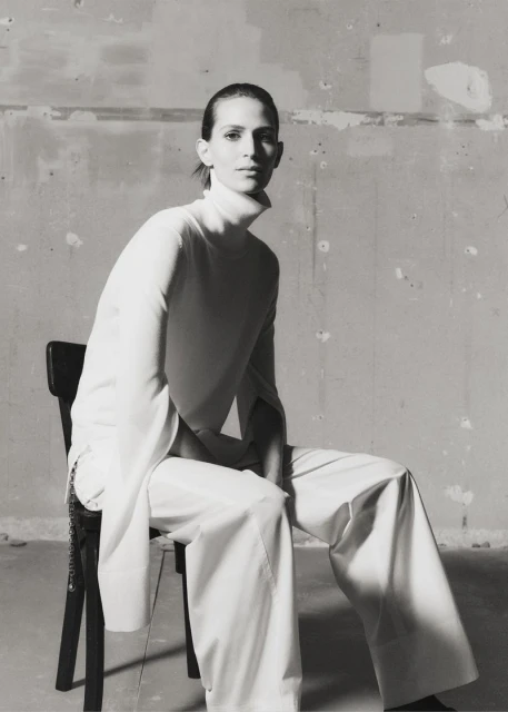 Model Jeanne Cadieu seated on a chair in a white turtleneck and trousers for Totême spring/summer 2025.