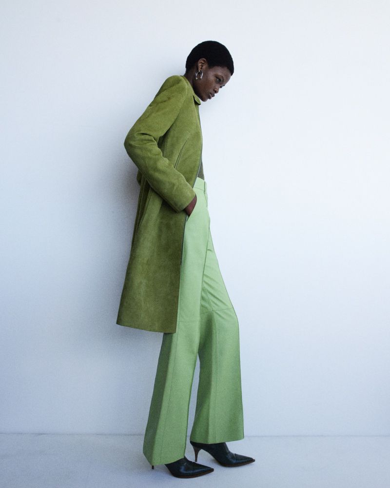 A woman poses in a studio wearing an olive green overcoat and light green slacks, showcasing modern fashion against a white background.