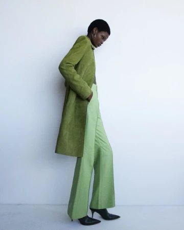 A woman poses in a studio wearing an olive green overcoat and light green slacks, showcasing modern fashion against a white background.
