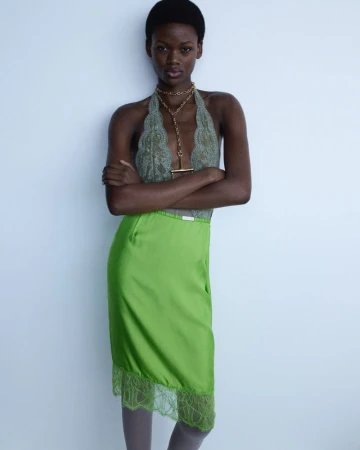 A slender woman poses in a lace halter top and green skirt, with her arms crossed, against a bright white backdrop in a fashion shot.