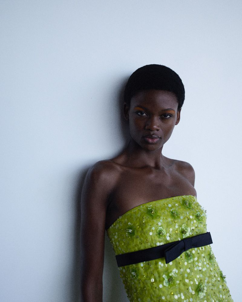 Black model Micklate Macobola wearing an embellished lime green strapless dress with a black bow against a pale studio wall.