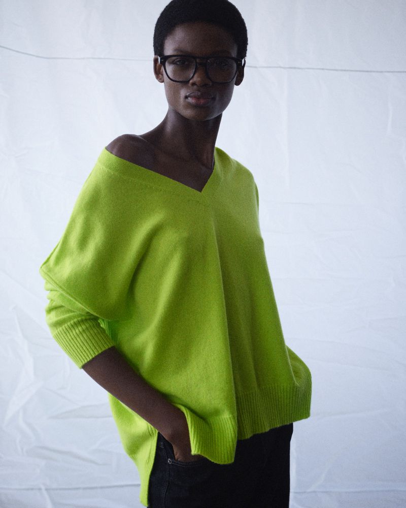 A Black female model poses in an off-the-shoulder lime green sweater and dark jeans against a white backdrop, showcasing a relaxed fashion style.