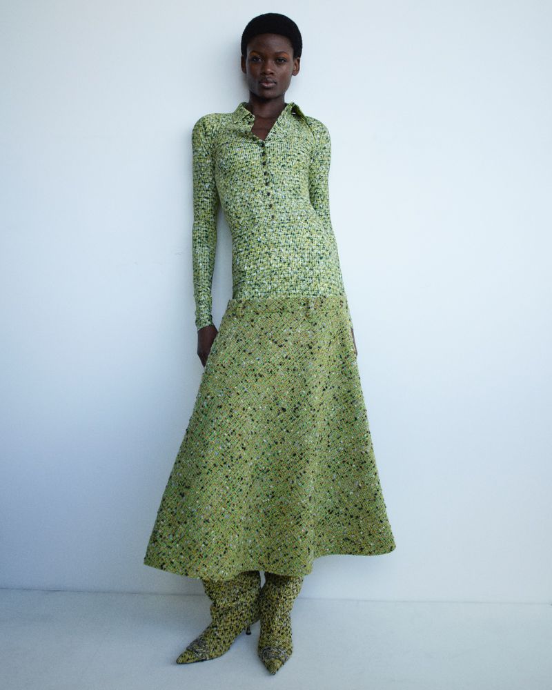 An adult female stands indoors against a white wall, wearing a green patterned dress and matching high-heeled boots in a full shot.