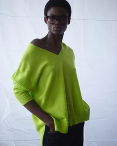 Model wearing black glasses and an oversized lime green V-neck knit sweater against a white draped backdrop.