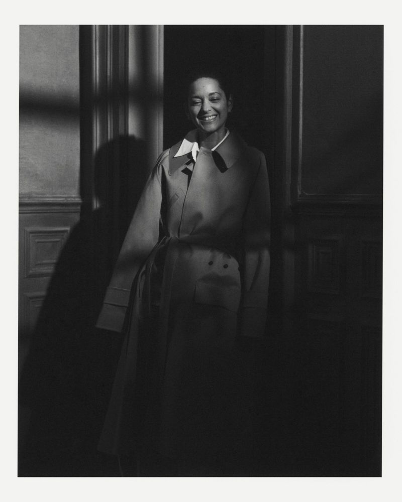 Marion Cotillard smiling in a belted trench coat in black and white portrait lighting against an interior doorway.
