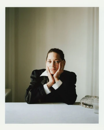 Portrait of a brunette woman in a black blazer at a white table, with hands on her face, and a neutral indoor background.