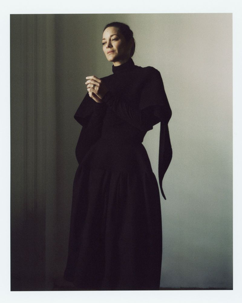 A woman poses in a long black gown with a high collar in a studio. The lighting is soft and creates a dramatic effect, highlighting her silhouette.