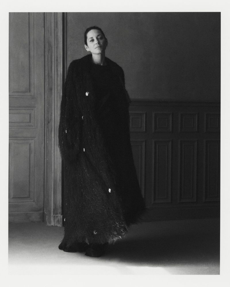 Portrait of an adult female fashion model wearing a long, dark embellished coat in an indoor setting with wall paneling.