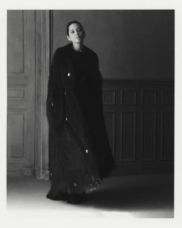 Portrait of an adult female fashion model wearing a long, dark embellished coat in an indoor setting with wall paneling.