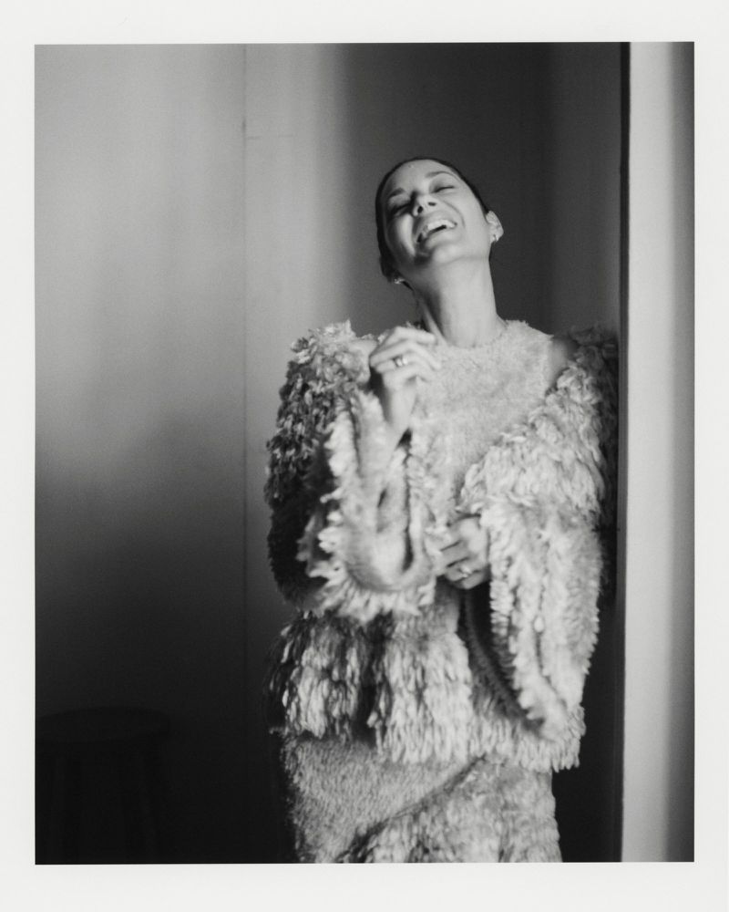 Monochrome portrait of a smiling woman in a textured outfit, captured indoors with soft lighting against a neutral wall backdrop.