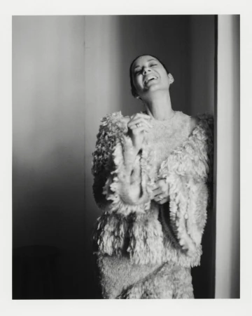 Monochrome portrait of a smiling woman in a textured outfit, captured indoors with soft lighting against a neutral wall backdrop.
