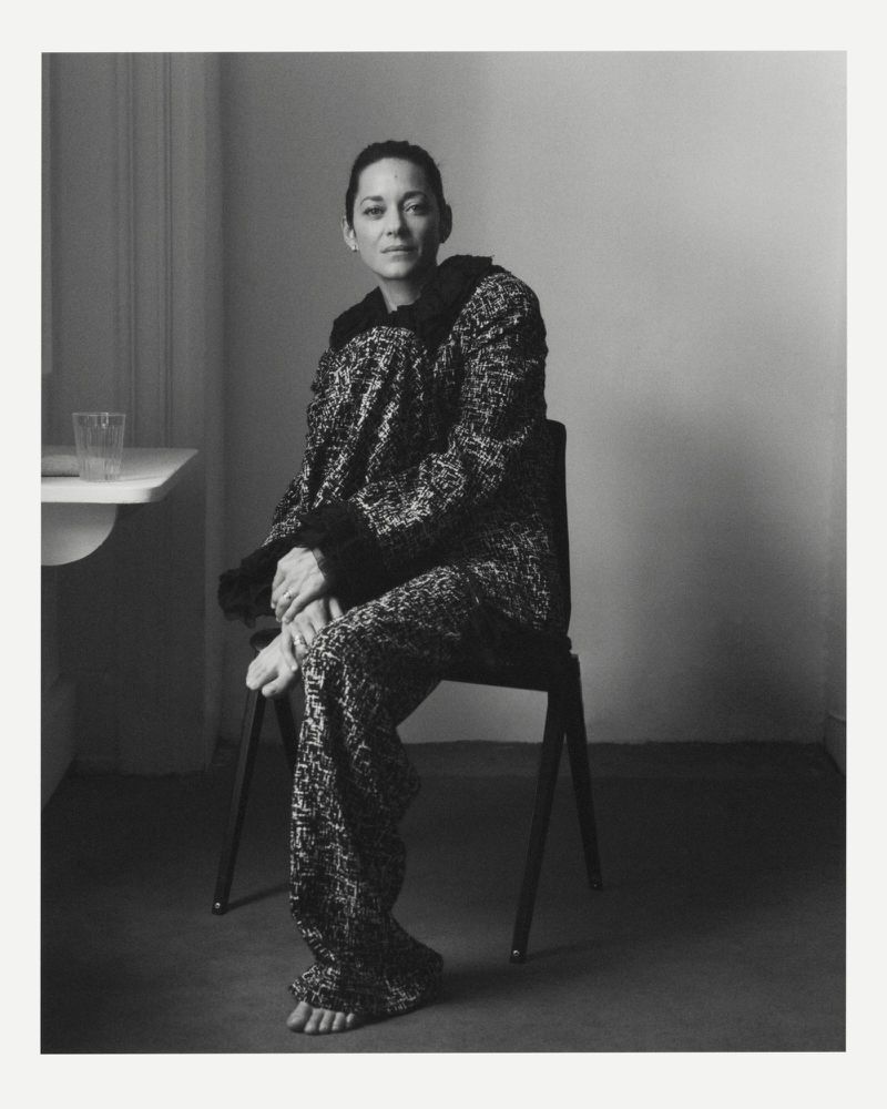 Monochrome portrait of a woman with dark hair wearing a patterned outfit, seated on a chair in an indoor setting near a small table.