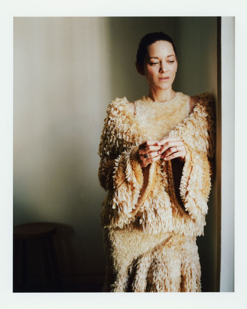 Portrait of Marion Cotillard in a textured, beige outfit against a neutral wall with soft lighting, with a stool visible in the background.