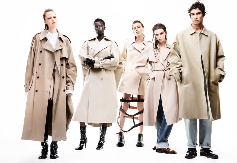 Fashion editorial shot of five diverse models wearing tan trench coats in an indoor photo studio with a white backdrop and studio lighting.