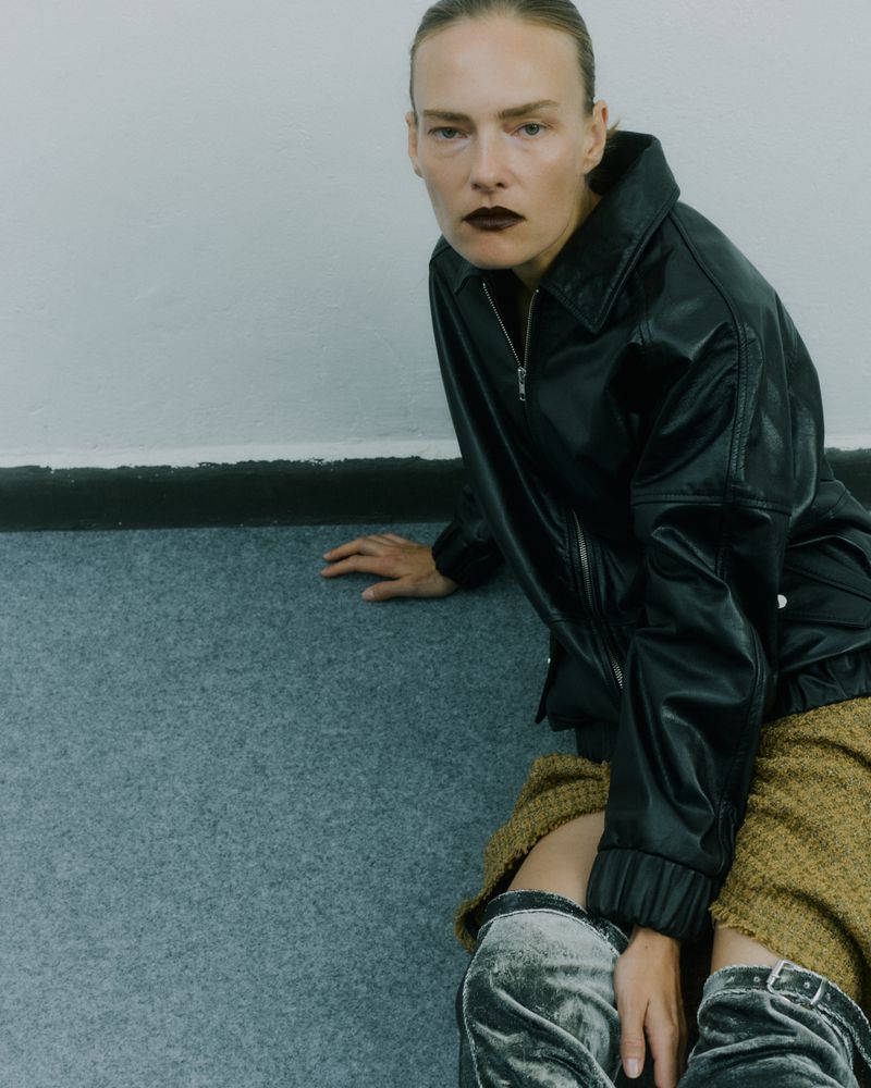 Adult woman wearing a black jacket and gray velvet boots sits on a blue carpet in front of a white wall in a studio shot.