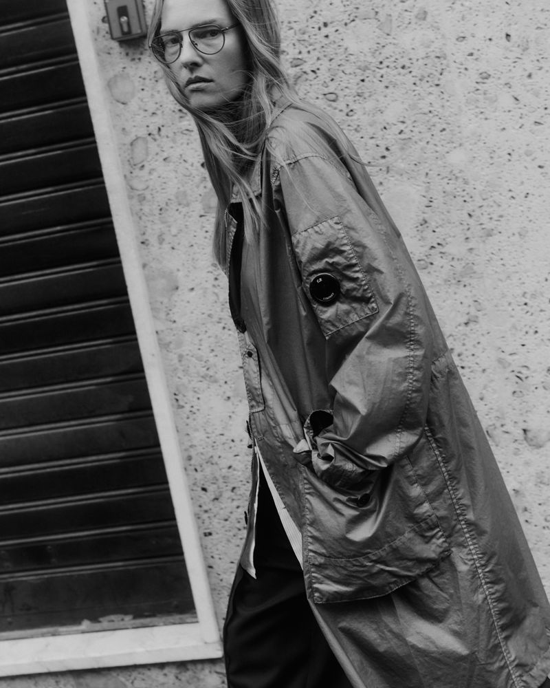 A stylish young adult woman with long blond hair wears a long olive-green jacket and glasses in an urban setting, shot in black and white.