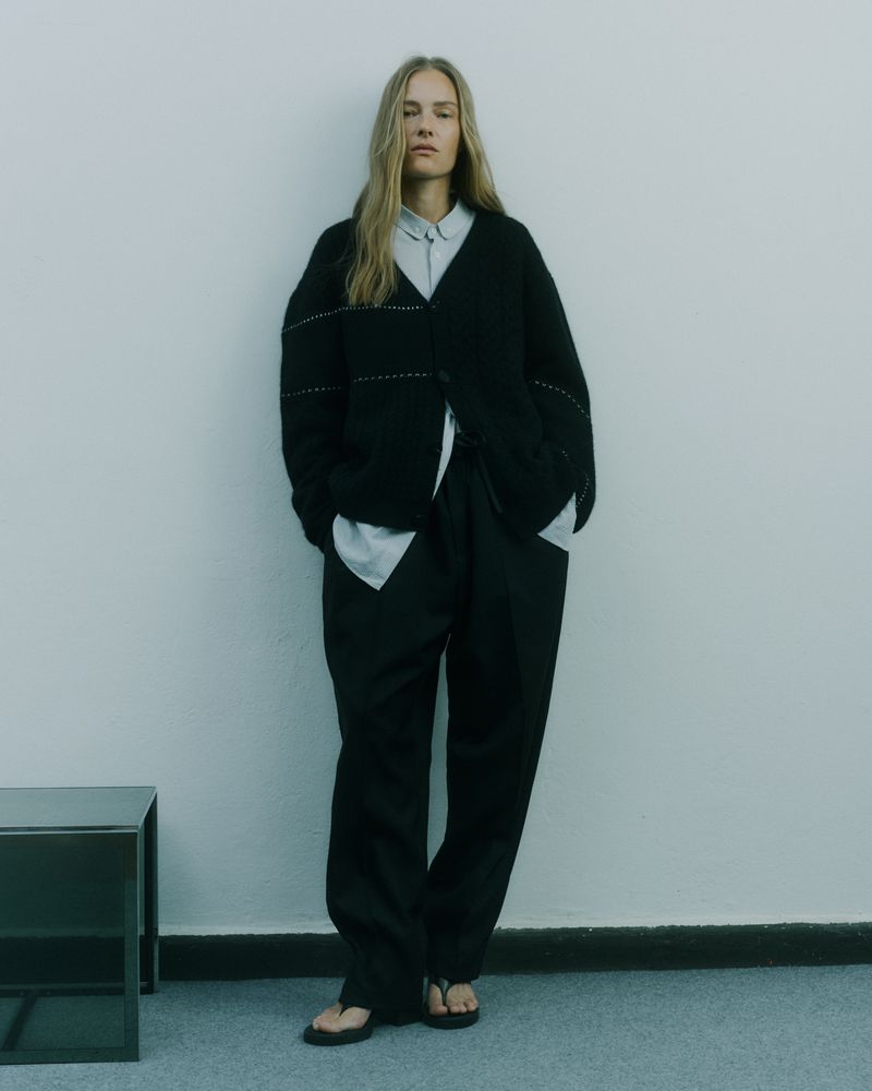 Fashion portrait of a woman in a black cardigan, light blue collared shirt, and black pants posing against a neutral background.