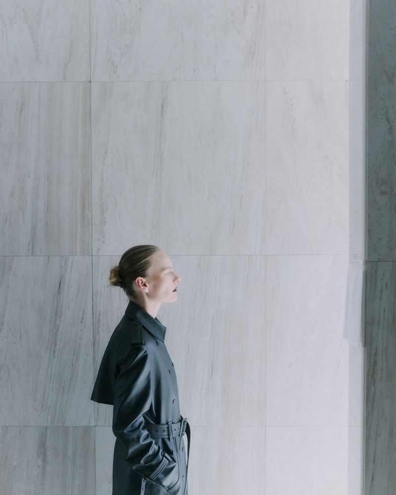 Woman in a stylish belted trench coat stands against a textured tile wall in a minimalist setting, bathed in soft, neutral lighting.