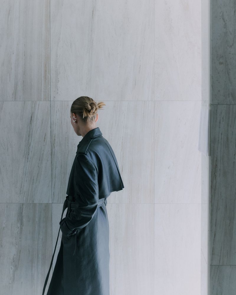 Blonde woman in a black trench coat stands against a textured white marble wall in a fashion or architectural context. Minimalist aesthetic.